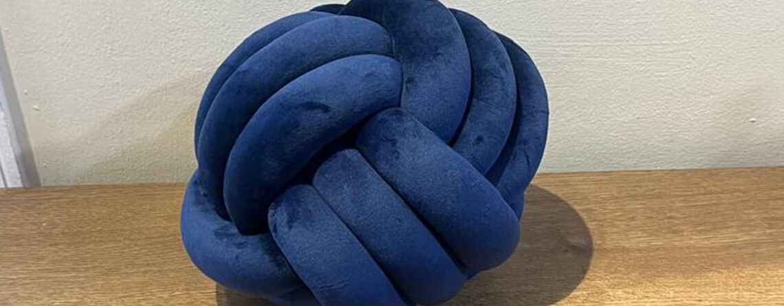 Blue knotted pillow made of suede fabric placed on a wooden surface.